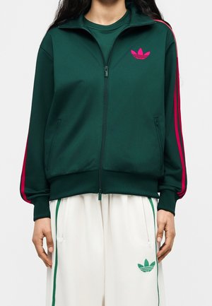 Person iført en mørkegrøn lynlåstrøje med pink striber på ærmerne og pink logo, kombineret med hvide bukser med grønne striber og grønt logo.