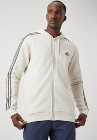 Sweatshirt cremosa com fecho, feita de um tecido suave, apresentando detalhes em cinza com três riscas ao longo das mangas e um logótipo no peito.