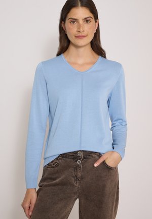 Femme aux longs cheveux bruns portant un pull bleu clair à col en V manches longues et un pantalon marron taille haute, une main dans la poche, fond neutre.