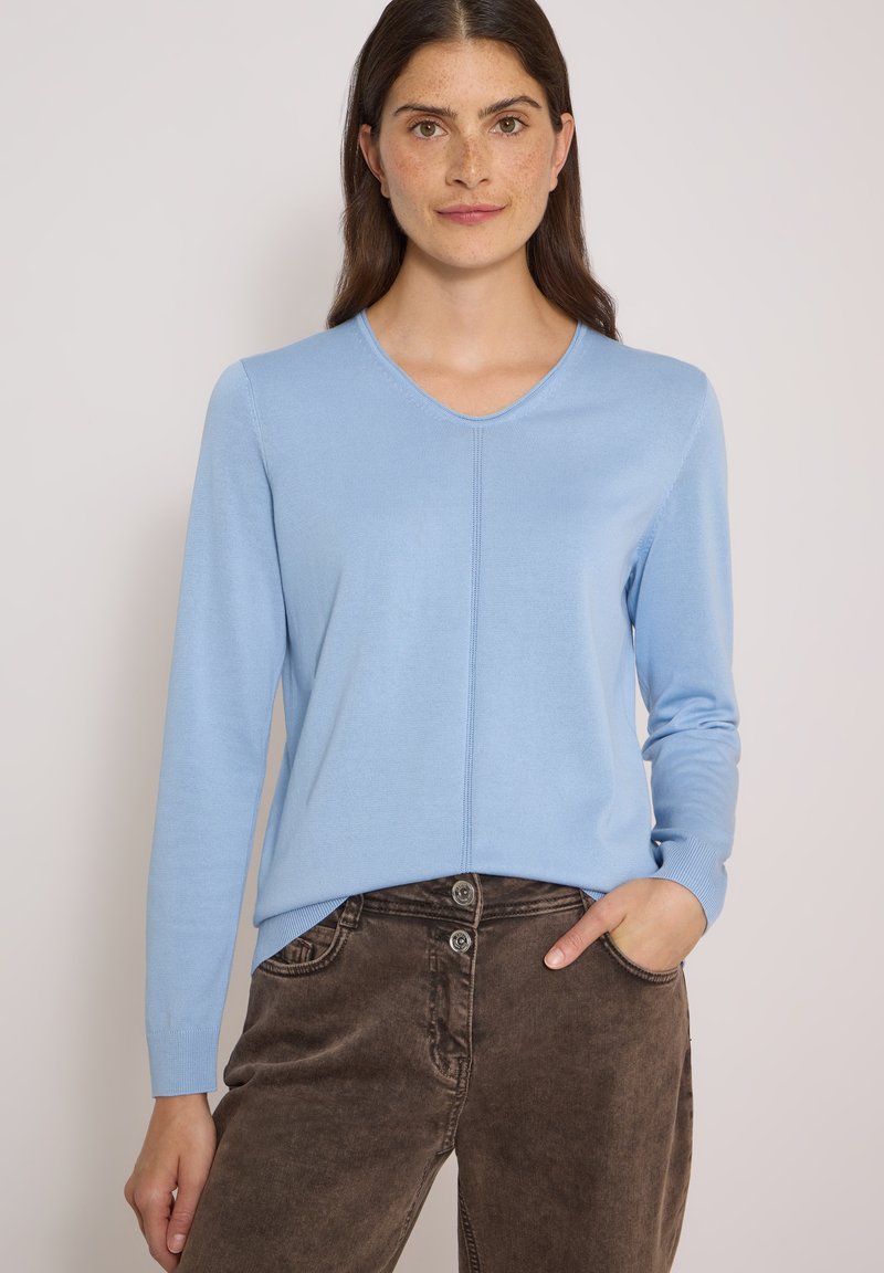 Femme aux longs cheveux bruns portant un pull bleu clair à col en V manches longues et un pantalon marron taille haute, une main dans la poche, fond neutre.