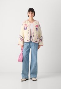 Polstret kremfarget jakke med fargerik brodering, rosa kant, brede ermer, paret med lyseblå jeans og en liten rosa håndveske.