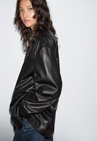 Zwarte leren jas met een relaxte pasvorm, opvallende textuur en opgerolde mouwen. Gecombineerd met donkere denim jeans, tegen een neutrale achtergrond.