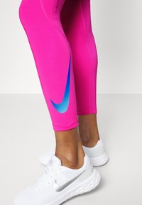 Leggings deportivos rosas con un swoosh azul de Nike, con un diseño ajustado y una textura suave, combinados con zapatillas blancas.