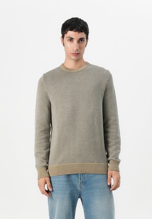 Knit sweater in beige with a textured grid pattern, round neckline, and ribbed cuffs. Paired with light blue denim jeans.