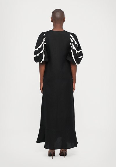 Rochie lungă neagră cu sleeves bufante, decorată cu dungi ondulate albe. Fusta este vaporoasă, iar materialul pare texturat. Este purtată cu tocuri maronii.