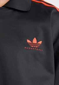 Fekete póló, textúrált anyagból; piros hímzett logóval rendelkezik, három levéllel és alatta a "BASKETBALL" felirattal. A galléron két piros csík található.