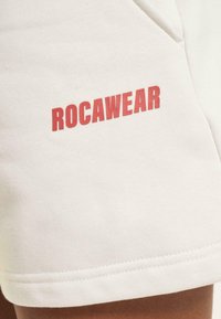 White cotton shorts featuring a red "ROCAWEAR" logo on the left side. Smooth texture with a straight cut and stitched hem.