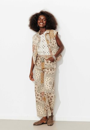 Femme avec coiffure afro portant un gilet et un pantalon à motifs, un haut en crochet blanc, des boucles d'oreilles créoles et des sandales, souriante, une main dans la poche.