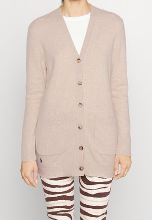 Cardigan beige con bottoni e tasche frontali indossato sopra una camicia bianca, abbinato a pantaloni striati in bianco e marrone a strisce zebra.