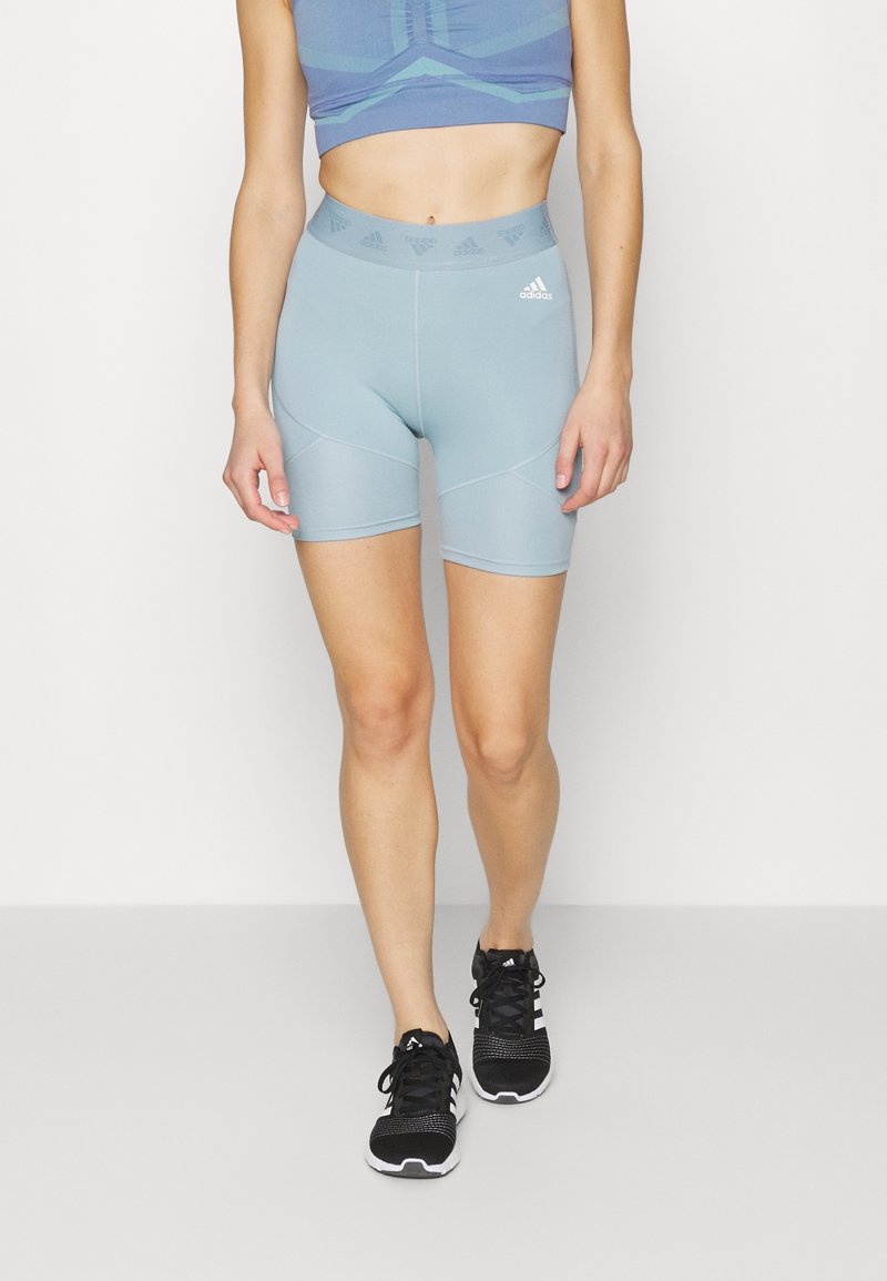 Femme portant un short de vélo taille haute bleu clair Adidas et un crop top assorti, marchant vers l'avant en baskets noires Adidas.