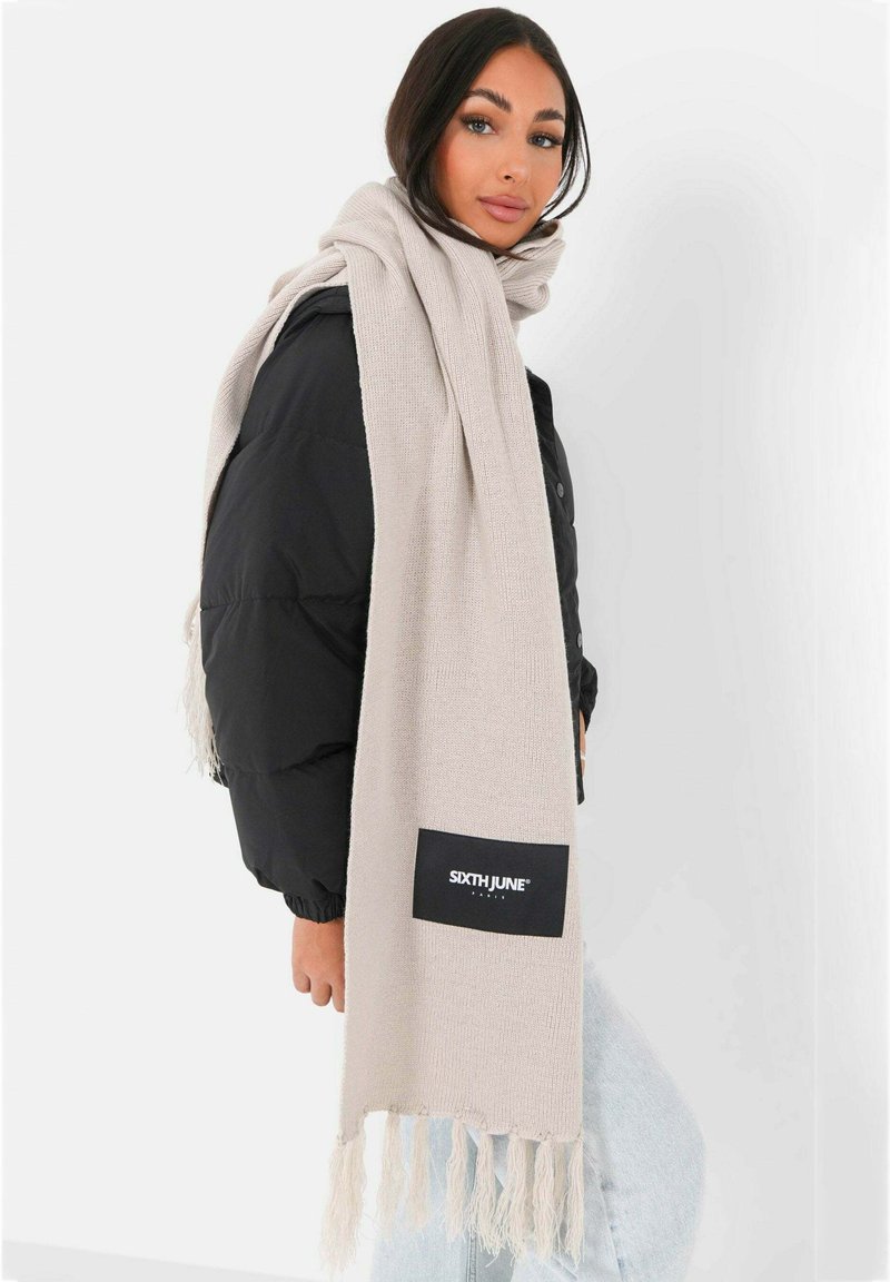 Beige gebreide sjaal met franjes en een zwart label met de tekst "SIXTH JUNE", over een zwarte pufferjas gedrapeerd.