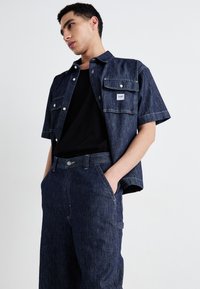 Dark denim short-sleeve shirt with front pockets, paired with dark denim pants. Features silver buttons and stitching accents.