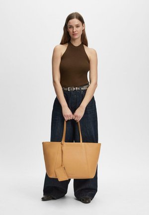 Frau in braunem ärmellosem Oberteil und dunkler Jeans mit weitem Bein, die eine große beigetöne Tote-Tasche mit kleinem angebrachtem Beutel hält, vor weißem Hintergrund stehend.