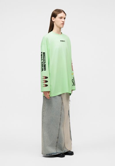 Άτομο με μακριά καστανά μαλλιά φοράει φαρδιά ανοιχτοπράσινη φούτερ με μαύρο και κόκκινο κείμενο και μακριά φούστα από πατς τζιν, μαζί με μαύρα παπούτσια.