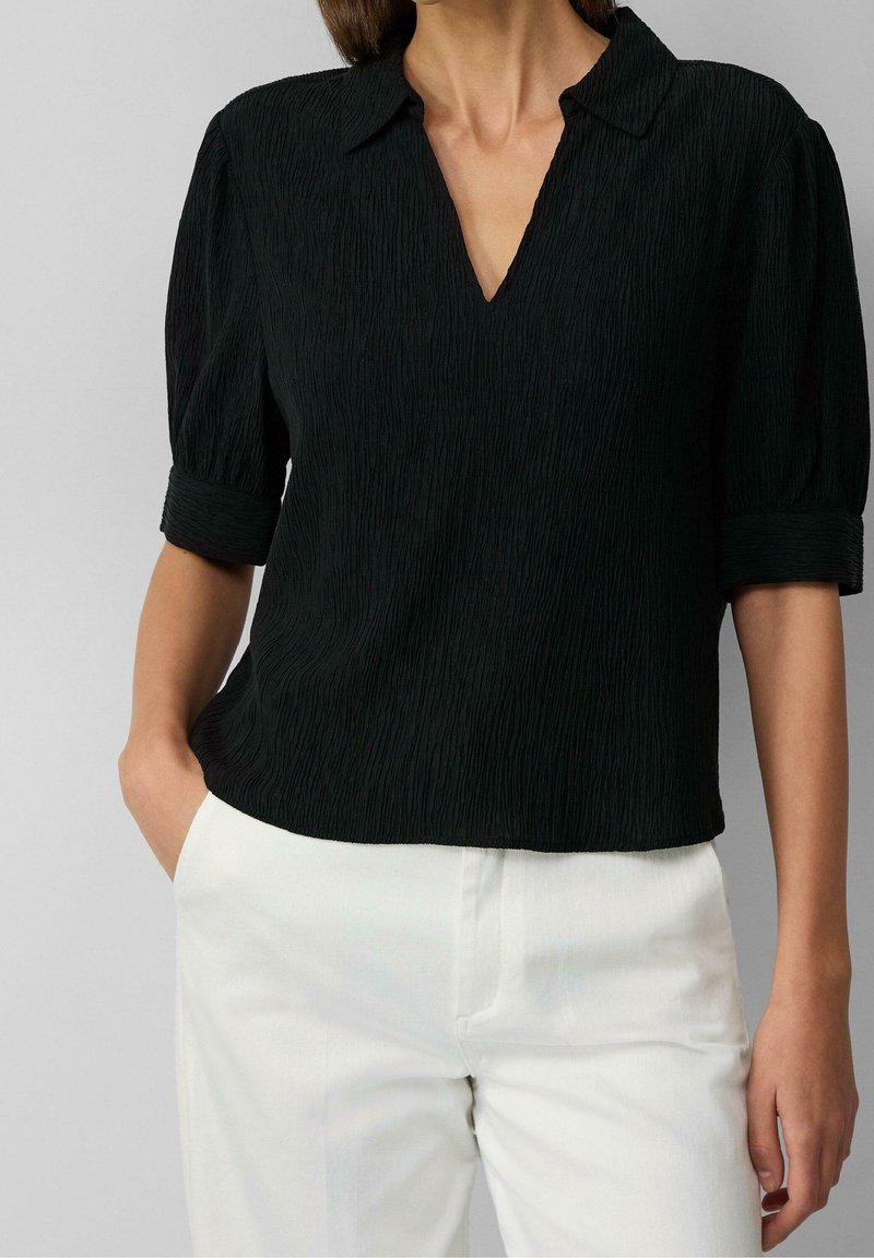 Femme portant un chemisier noir texturé avec des manches trois-quarts retroussées et un pantalon blanc, une main dans la poche sur un fond neutre.