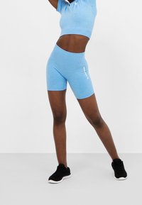 Dospělý muž v modrých be seam workout šortkách a topu, stojící s rukama vztyčenýma, s černými sportovními botami na jednolité šedé podlaze.