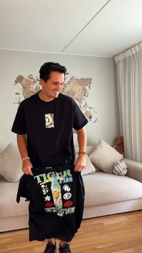 Smilende mann i svart T-skjorte som holder en annen svart T-skjorte med fargerik "TIGER" design, står i stuen med verdenskart som veggkunst.