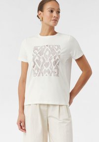 Biała bawełniana koszulka z osobliwym, teksturowanym wzorem geometrycznym w subtelnym odcieniu beżu. Okrągły dekolt i krótkie rękawy zapewniają luźny krój.