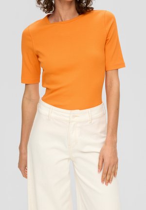 Žena v oranžovém přiléhavém tričku s krátkým rukávem zastrčeném do kalhot s vysokým pasem a širokými nohavicemi v bílé barvě na jednobarevném pozadí.