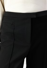 Černé kalhoty na míru mají hladkou texturu, slim fit a nenápadnou boční kapsu s malým zipem. Pás je plochý.