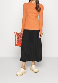 Ribbad orange långärmad topp; svart plisserad midi kjol; röd stickad väska; beige träskor med trädsål och silverdetaljer.