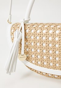 Vävd beige väska med vita detaljer, utrustad med en tofs och en guldfärgad logotypbricka. Rund form med texturerad yta och kontrasterande färger.