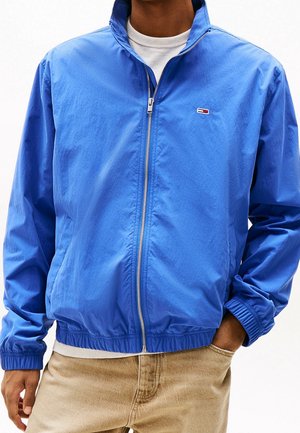 Mann trägt eine leuchtend blaue Zip-up-Jacke über einem weißen Hemd und beigen Hosen, steht mit einer Hand in der Tasche vor weißem Hintergrund.