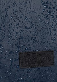 Marineblauw waterdicht stof met regendruppels en een getextureerd oppervlak; voorzien van een zwarte, geperste logo-label met de tekst "EASTPAK".