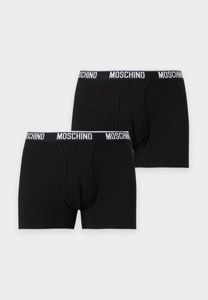 Un mannequin masculin se tient devant un fond de studio gris clair, vêtu d’un boxer noir Moschino.