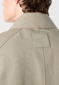 Lichtbeige jack met een gestructureerde kraag, voorzien van een vierkante groene leren logo-patch en zichtbare stiksels op de achterkant.