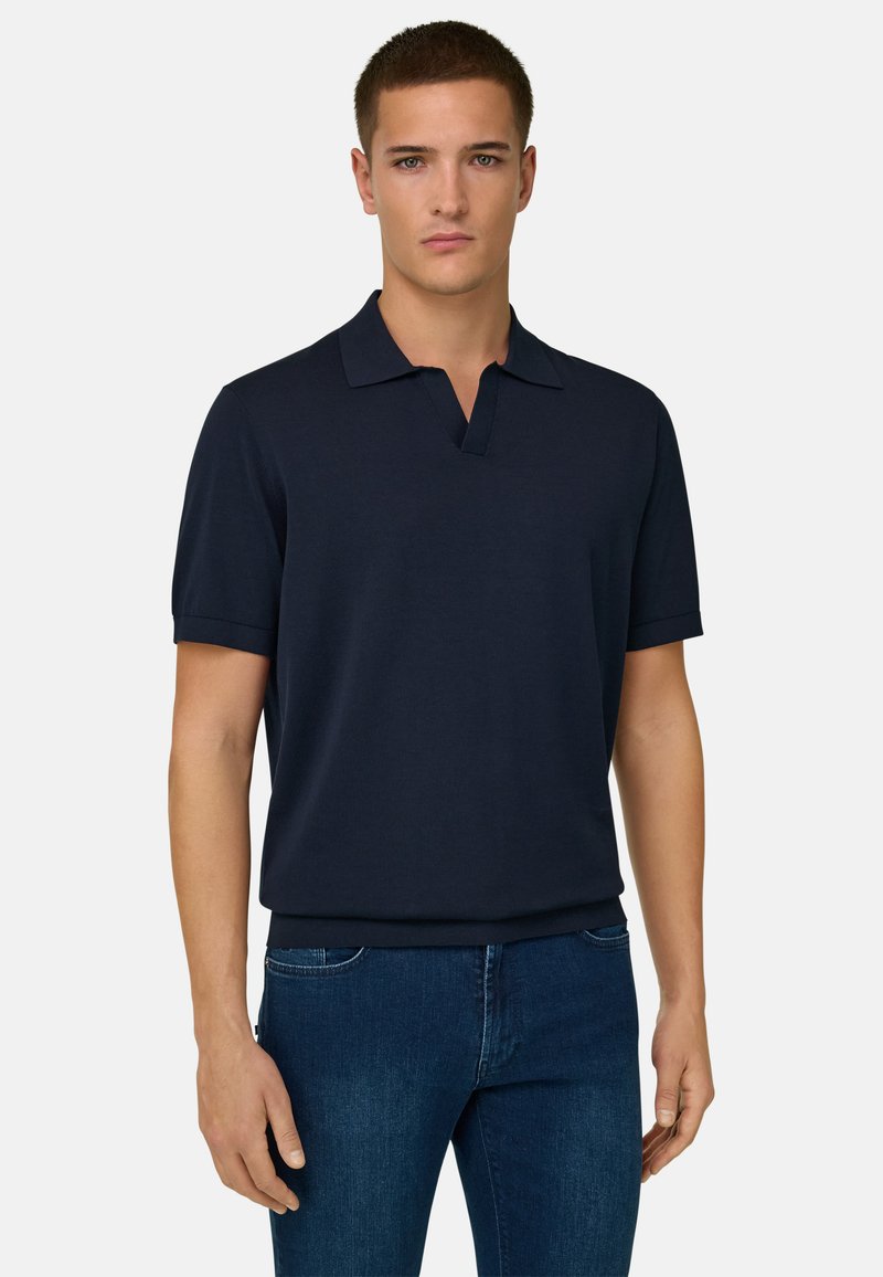 Homme portant un polo bleu marine à manches courtes et un jean bleu, debout devant un fond uni gris clair.