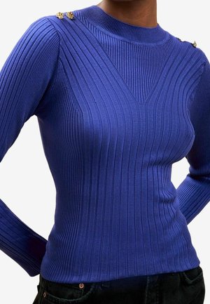 Gros plan d'une personne portant un pull violet ajusté à côtes, manches longues, avec des boutons dorés sur les épaules et un pantalon noir.