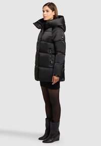 Mustast värvi puffer-jope kõrge kaeluse, tikandmustri, külgse tõmblukuga tasku ja veekindlast kangast. Pähe on tõmmatud must kampsunkleit.