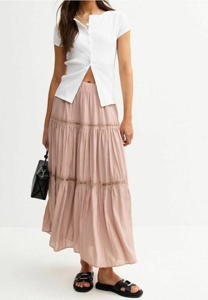 Femme portant un haut blanc à manches courtes avec boutons, une longue jupe légère rose à volants avec bordure en dentelle, des sandales noires et un sac à main noir.