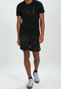 Camiseta deportiva negra con la frase "JUST DO IT" en acabado mate, combinada con pantalones cortos negros con estampado y zapatillas Nike grises con detalles negros.