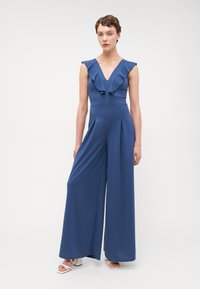 Macacão azul com decote em V franzido, pernas largas e detalhes plissados. Feito de tecido suave; usado com sandálias de salto bloco brancas.