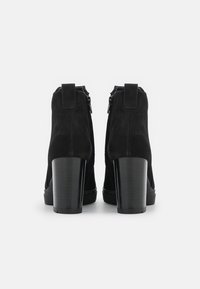 Para czarnych zamszowych botków za kostkę z wysokimi słupkowymi obcasami, przedstawionych od tyłu na jednolitym białym tle.