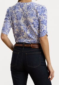 Kvinna som bär en blå blommönstrad topp med ärmar som når till armbågarna, instoppad i mörkblå jeans med ett brunt bälte, visad från ryggen.