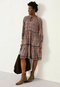 Langärmliges, gestuftes Kleid mit floralem Muster in Pink, Marineblau und Creme. Braune Knöchelstiefel und eine dunkelbraune Umhängetasche runden den Look ab.
