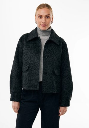 Vrouw draagt een zwarte jas met luipaardprint en knopen over een grijze coltrui en zwarte broek, staand voor een witte achtergrond.
