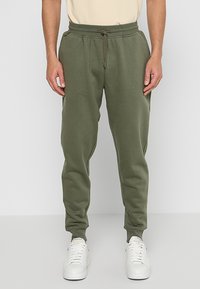 Pantalones de chándal verdes con cintura elástica y cordón. Cuenta con tobillos ajustados y una textura suave. Llevados con zapatillas blancas.