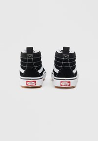 Mustat korkeat tennarit, joissa on valkoiset raidat, mokkanahkaa, valkoinen pohja ja punainen Vans-logo takana. Varustettu teksturoidulla kumipohjalla.