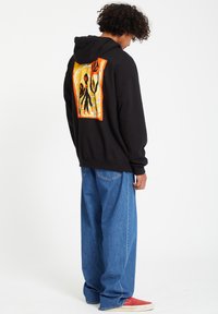 Zwarte hoodie met een kleurrijk abstract ontwerp op de achterkant. Gecombineerd met losse blauwe jeans en rode schoenen, wat een casual streetwear-stijl laat zien.