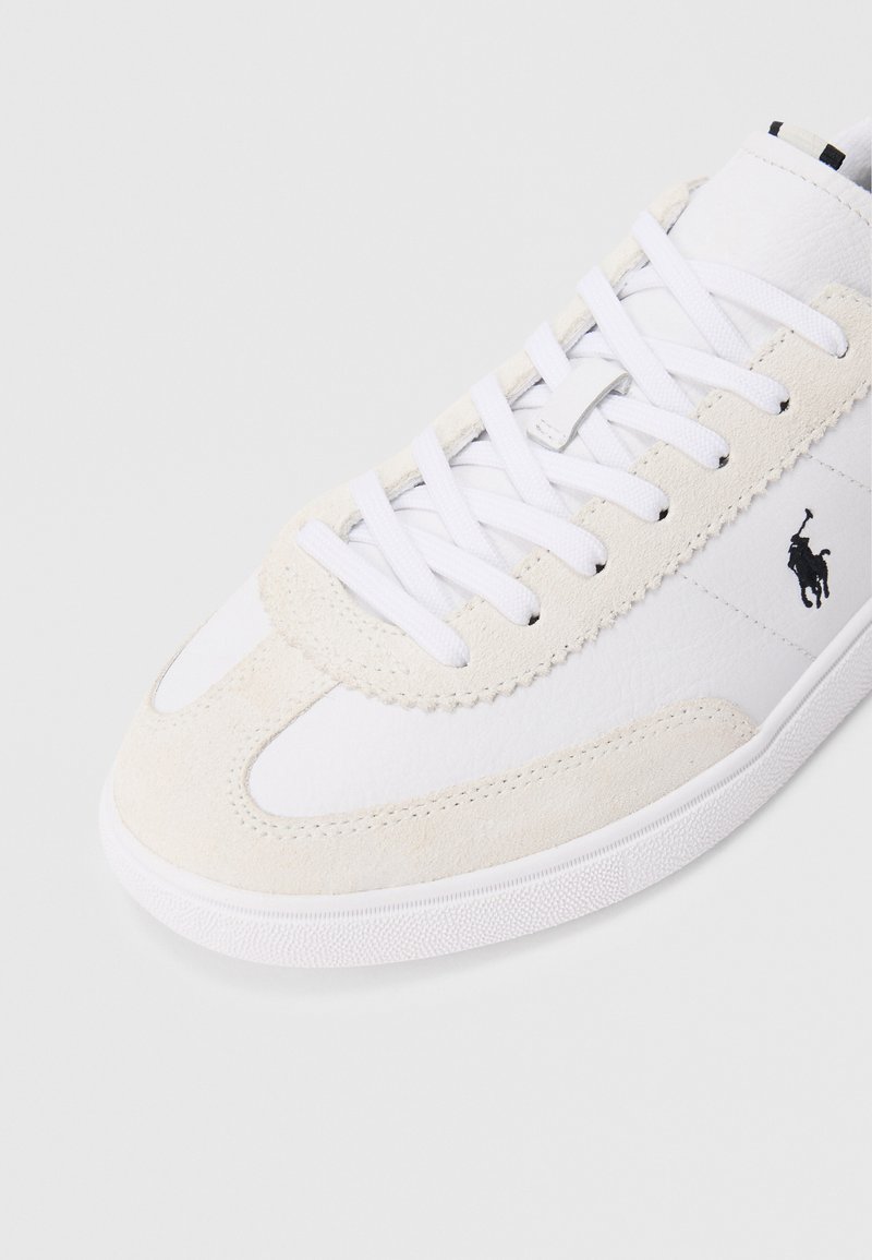 Sneaker bianca con dettagli in suede e lacci bianchi, caratterizzata da un logo di un giocatore di polo nero sul lato e una suola in gomma con texture.