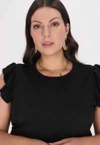 Sort top lavet af blødt stof, med puffede ærmer. Accessoriseret med lagdelte guldhalskæder og store guldringe.