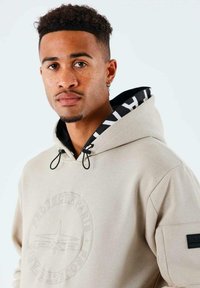 Hoodie beige avec logo brodé, accents noirs, cordons de serrage réglables et capuche intérieure à motif. Fabriqué en tissu doux et épais.