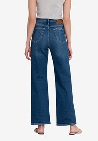 Achteraanzicht van een persoon die hoge, wijde blauwe jeans, een witte mouwloze top en beige geweven instappers draagt op een witte achtergrond.