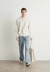 Λευκό πουκάμισο με μακριά μανίκια και γιακά, ανοιχτό μπλε παντελόνι ίσιας γραμμής και μαύρα αθλητικά παπούτσια. Κρατάει μια μπεζ τσάντα.