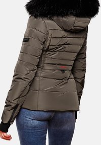 Vrouw in een gewatteerde olijfgroene jas met een zwarte bontkraag en blauwe jeans, van achteren gezien tegen een effen achtergrond.