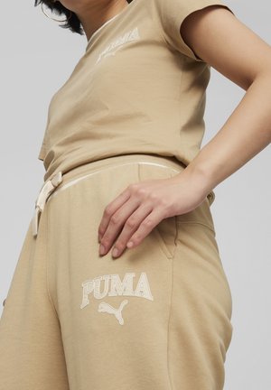 Camiseta de algodón beige y pantalones de chándal con cinturilla elástica. La camiseta presenta un logo bordado en blanco de "PUMA"; los pantalones tienen detalles del logo y cordón.